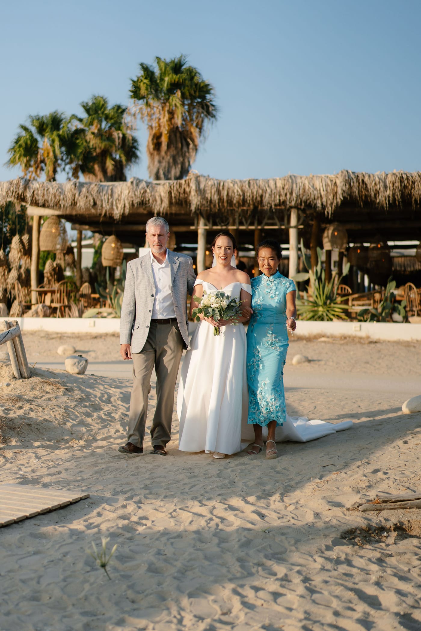 The bride walking down the aisle with her parents on the sand